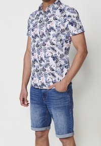 Homme portant une chemise blanche à manches courtes avec un imprimé de feuilles bleues et roses, associée à un short en jean bleu retroussé, debout devant un fond clair.