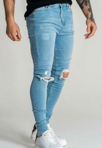 Jeans ajustados de mezclilla azul claros con roturas, combinados con zapatillas blancas, parte inferior del torso y piernas visibles, brazos tatuados a los lados.