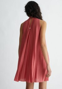 Donna con lunghi capelli scuri che indossa un vestito rosa antico lucido senza maniche e plissettato, vista da dietro, in piedi contro uno sfondo chiaro e uniforme.