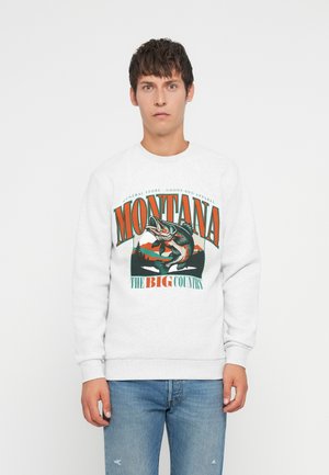 Jonge man in een wit sweatshirt met een kleurrafische afbeelding van vissen in Montana en een blauwe jeans, staande tegen een effen witte achtergrond.