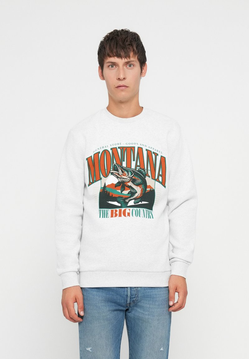 Jeune homme portant un sweat-shirt blanc avec un graphique de pêche coloré du Montana et un jean bleu, se tenant devant un fond blanc uni.