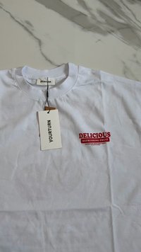 T-shirt de algodão branco com decote redondo, apresentando texto impresso em vermelho "FRUTA REFRESHING DELICIOSA" e uma etiqueta anexada com o nome da marca "YOURTURN."