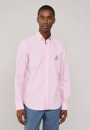 Camicia a righe rosa chiaro con maniche lunghe, colletto button-down, bottoni bianchi e un piccolo logo ricamato sul petto. Indossata con pantaloni blu.
