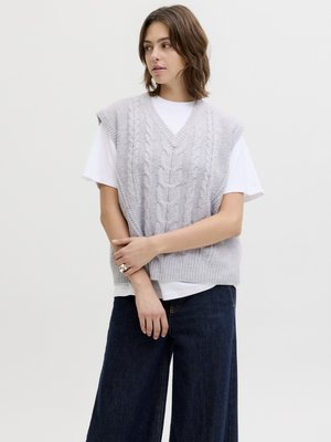 Kvinde med skulderlangt brunt hår iført en lysegrå kabelstrikket vest over en hvid T-shirt og mørke vide jeans, der står med armene krydset.