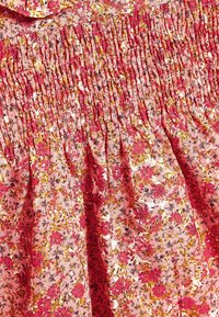 Detail květinových šatů s nařasenou, texturovanou růžovou látkou s drobnými květinami v červené, žluté a černé barvě. Jemné, delikátní vzory po celém povrchu.