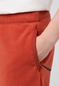 Roestkleurige fleece broek met een zijzak, voorzien van een decoratieve zwarte trekkoord en afwerking details. Zachte textuur en casual ontwerp.