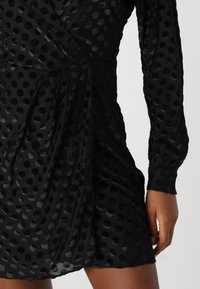 Robe noire à pois en tissu texturé, présentant un design croisé, des manches longues et une coupe légèrement drapée.