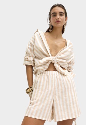 Frau trägt beige-weiß vertikal gestreifte Shorts und dazugehöriges lockeres Hemd, das in der Taille gebunden ist, mit großen sonnenförmigen Ohrringen und Armbändern als Accessoires.