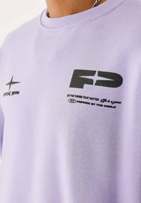 Camiseta de algodón lila con estampados gráficos en negro: un logo de estrella, "PXP" y "FD" con texto abajo. Textura suave y escote redondo.