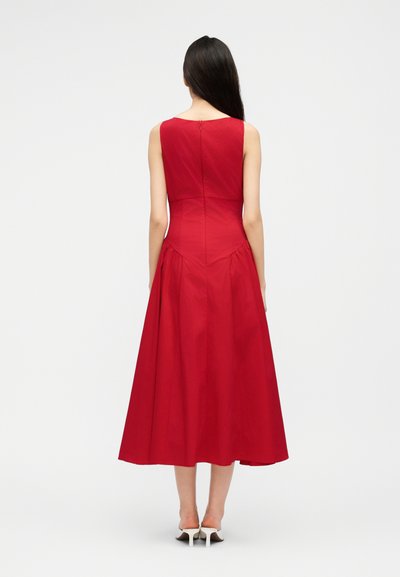 Γυναίκα με μακριά σκούρα μαλλιά, ντυμένη με αμάνικο κόκκινο φόρεμα μήκους μίντι και λευκές γόβες, όρθια με την πλάτη προς το φακό, μπροστά σε απλό ανοιχτόχρωμο φόντο.