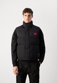Crni утепљени prsluk s visokim ovratnikom, sa crvenom oznakom. Dizajn uključuje velike prednje džepove i elegantnu quilted teksturu.