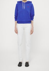 Blaues Baumwoll-Hoodie mit Kordelzug, Kängurutasche und rotem Logo; kombiniert mit weißen Jeans und schwarzen Loafers.