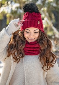 Berretto lavorato a maglia rosso con un pompon in pelliccia nera, abbinato a una sciarpa lavorata a maglia coordinata. Il modello indossa una giacca in pile color crema e un maglione chiaro.