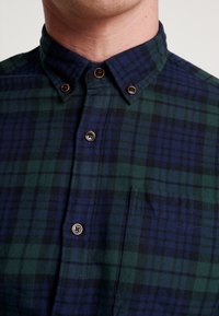 Camicia a quadri verde e blu navy con colletto, caratterizzata da dettagli con bottoni marroni e una tasca sul petto. Tessuto morbido.