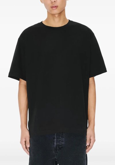 Person iført en ensfarvet sort oversized t-shirt med korte ærmer og mørke jeans, stående mod en ensfarvet hvid baggrund.