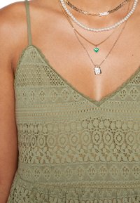 Detailní záběr na osobu v zelených krajkových šatech s tenkými ramínky a vrstvami náhrdelníků, které obsahují perly, zelené srdce a přívěsek ve tvaru zámku.