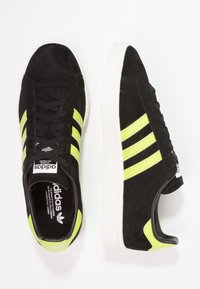 Svarta mockaskor med tre djärva neon-gula ränder och en vit gummisula, samt en häl i svart och gult.