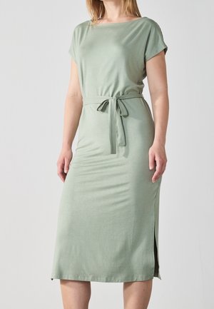 Donna che indossa un abito midi verde chiaro a maniche corte con cintura in vita e spacco laterale, in piedi davanti a uno sfondo semplice.