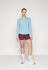 Hellblaues Langarmoberteil kombiniert mit burgunderfarbenen Shorts. Das Modell trägt weiße Knöchelsocken und hellere Sneakers. Einfaches, sportliches Design.