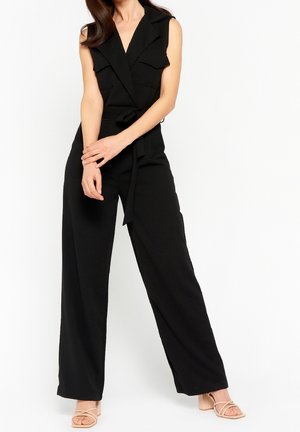 Femme portant une combinaison noire sans manches, jambes larges et ceinture nouée, posant avec les bras croisés sur fond blanc.