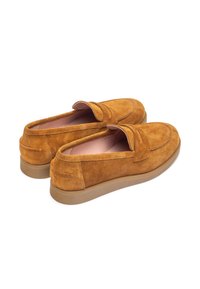 Mocasines de ante marrón con una textura suave, suela de goma beige de perfil bajo y un diseño costurado a lo largo de la parte superior. Sin logotipos ni adornos visibles.