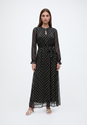 Femme debout portant une longue robe noire à pois avec des manches transparentes, une ceinture à la taille et des sandales blanches à talons.