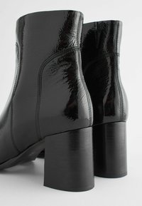 Bottes noires à la cheville en cuir verni brillant avec une texture lisse, dotées d'un talon bloc et de coutures renforcées.