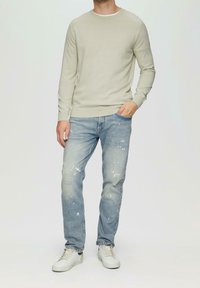 Hellgrauer Strickpullover, blaue Jeans im Used-Look mit Farbspritzern und weiße Turnschuhe. Einfaches Rundhalsdesign und schmaler Schnitt.