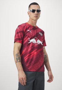 Maglia atletica rossa e marrone con un design a gradiente, caratterizzata da loghi bianchi e un colletto girocollo. Realizzata in tessuto liscio e leggero.