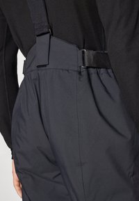 Crne vanjske hlače imaju podesive bretele, elegantan, glatak materijal s mat finišem i pojas za sigurno prianjanje.
