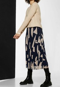 Maglione a maglia beige con maniche lunghe abbinato a una gonna a pieghe blu navy con un motivo geometrico beige. Stivaletti neri alla caviglia completano il look.