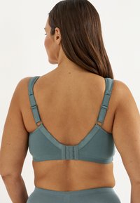 Frau mit langen braunen Haaren, von hinten zu sehen, trägt einen verstellbaren BH mit Trägern in Teal und passende hochtaillierte Unterteile vor einfachem Hintergrund.