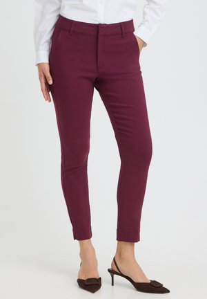Femme portant un pantalon slim rouge foncé, une chemise blanche et des escarpins noirs à bout pointu avec les talons relevés.