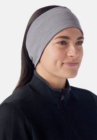 Une femme aux longs cheveux noirs portant un bandeau en tissu gris et une veste zippée noire, souriant légèrement, de profil à droite sur un fond blanc.
