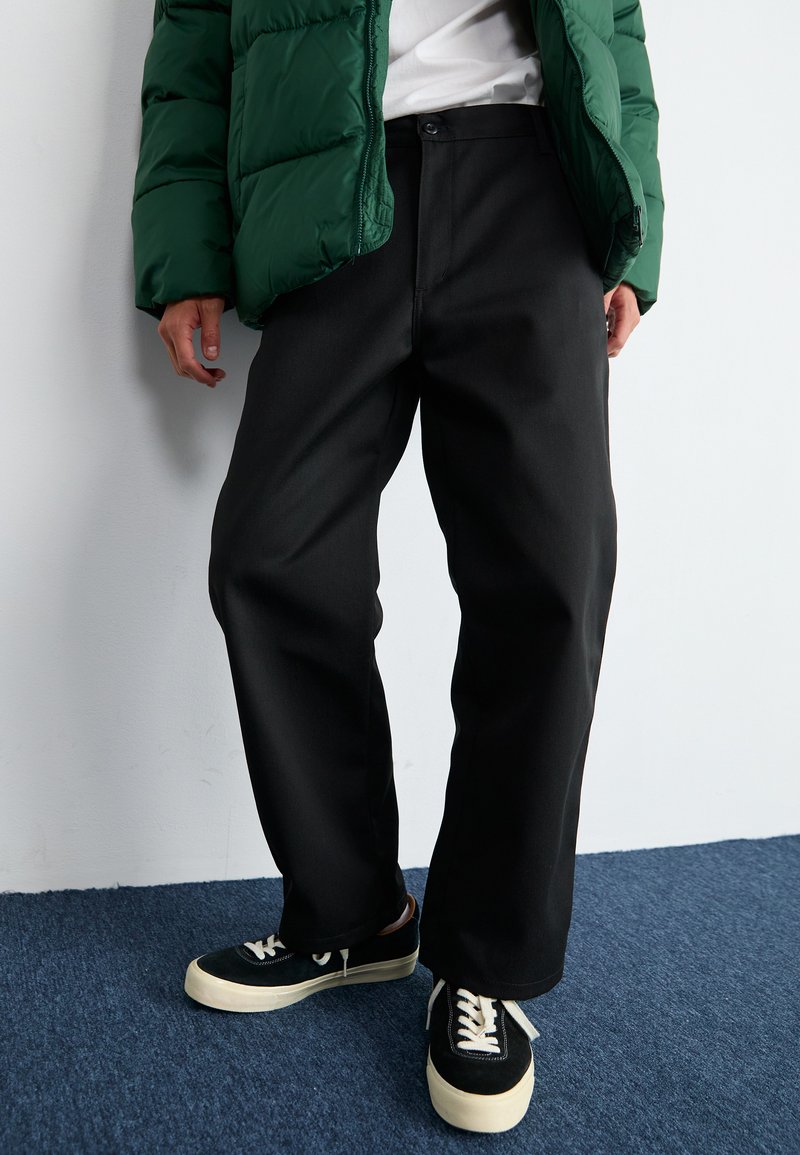 Pantaloni neri a gamba dritta realizzati in un tessuto liscio, con chiusura a bottone e vestibilità comoda. Indossati con un giubbotto imbottito verde e sneakers.
