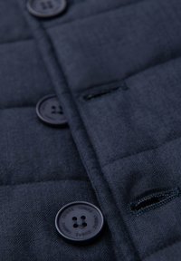 Detail tmavě modré prošívané látky s černými knoflíky označenými "OMBRE" a prošitými dírkami na knoflíky.