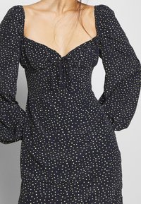 Marineblaues Kleid mit puffärmel, das eine Schleifenfront und gelbe Punkte aufweist. Glatter Stoff mit tailliertem Schnitt.