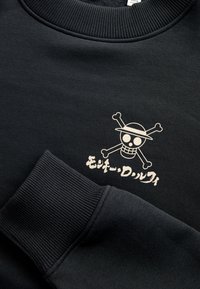 Sort sweatshirt med ribbet rund halsudskæring og ærmekanter. Den har et hvidt trykt motiv af en dødningehoved med korslagte ben og tekst nedenunder.