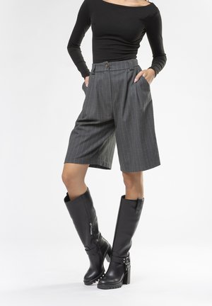 Shorts grigi a righe con vita alta e tasche laterali, abbinati a stivali neri in pelle con tacco al ginocchio, decorati con borchie.