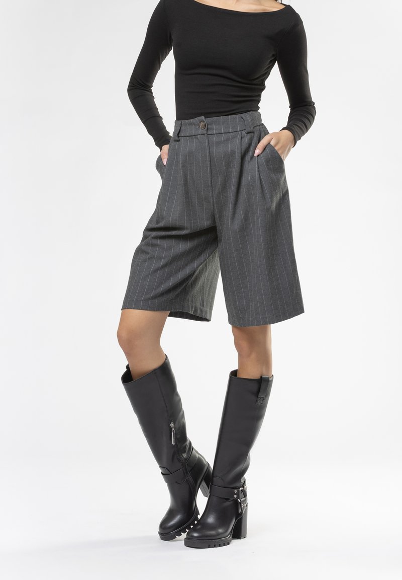 Shorts grigi a righe con vita alta e tasche laterali, abbinati a stivali neri in pelle con tacco al ginocchio, decorati con borchie.