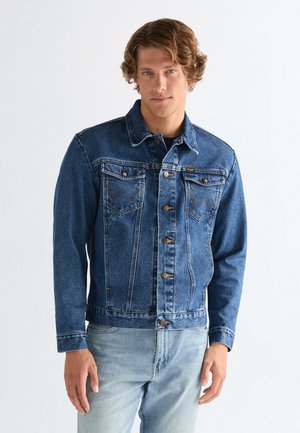 Hombre con cabello ondulado, vestido con una chaqueta vaquera azul abotonada y vaqueros azul claro, de pie contra un fondo liso y claro.