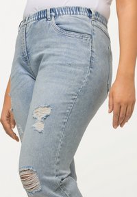 Jeans de mezclilla azul claro con un corte relajado, que presentan detalles desgastados, parches deshilachados y una cinturilla elástica para mayor comodidad.