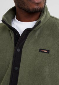 Fleece jacket in olive green with a black button placket and collar. Features a rectangular logo patch reading "TOG24" in orange.