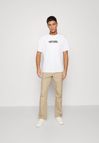 Weißes Baumwoll-T-Shirt mit dem Aufdruck "VIRTUAL" in Schwarz, kombiniert mit hellbeigen Baumwollhosen und weißen Sneakers. Einfaches, lässiges Design.