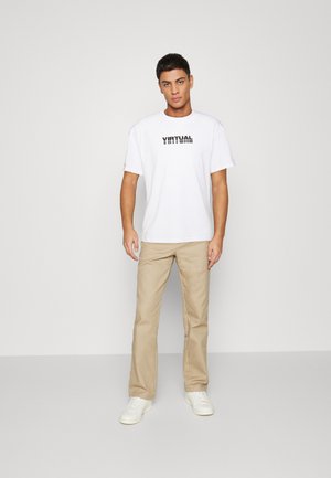 Weißes Baumwoll-T-Shirt mit dem Aufdruck "VIRTUAL" in Schwarz, kombiniert mit hellbeigen Baumwollhosen und weißen Sneakers. Einfaches, lässiges Design.