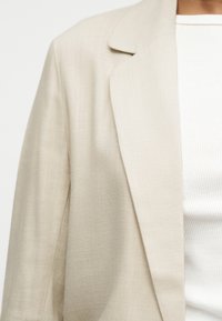 Μπεζ υφασμάτινο σακάκι φορεμένο πάνω από λευκό ριπ πλεκτό πουκάμισο, κοντοκομμένο για να δείχνει τον ώμο και το πάνω μέρος του στήθους.