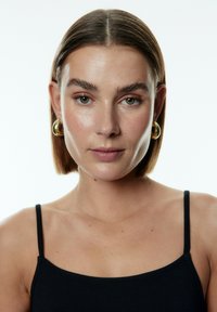 Schwarzes ärmelloses Top mit dünnen Trägern; glatte Textur; das Model hat kurze braune Haare und trägt goldene Creolen; neutraler Hintergrund.