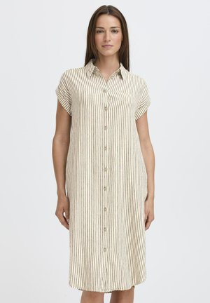 Nő, aki bézs színű, vékony fekete függőleges csíkokkal díszített, gombos, rövid, felhajtott ujjú ruhát visel, és egy egyszerű háttér előtt áll.