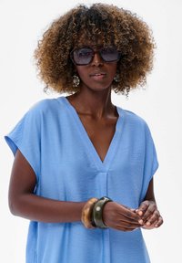 Jasnoniebieska lniana bluzka z dekoltem w serek i krótkimi rękawami, zestawiona z dużymi ciemnymi okularami przeciwsłonecznymi oraz bransoletkami z żywicy w różnych kolorach.