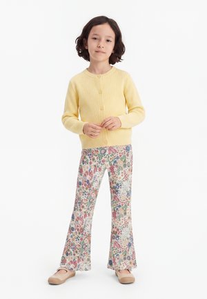 Pige med mørkt hår har en gul knappet cardigan på, bukser med blomsterprint og beige sko, og står foran en hvid baggrund.